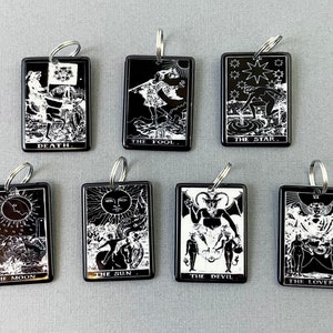 May include: A collection of black rectangular keychains featuring white illustrations of tarot card designs. Each keychain has a silver keyring and displays a different card, including "Death," "The Fool," and "The Star."