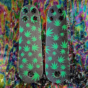May include: Two skateboard decks with a black background and a repeating pattern of green cannabis leaves. Each deck has four black mounting holes with red accents and a circular hole at the bottom.