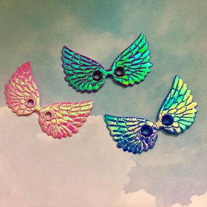 May include: Three iridescent wing-shaped embellishments in pink, green, and blue. Each wing has a central hole and a textured, feathered design. The wings are set against a cloudy, light blue background.