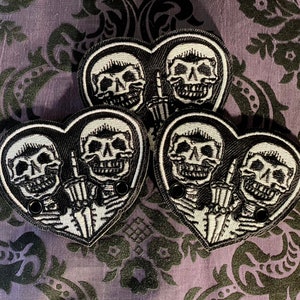 May include: Heart-shaped black and white embroidered patches featuring two skeleton figures. One skeleton is giving the middle finger. The patches have a black border and two black grommets. The background is a patterned fabric.