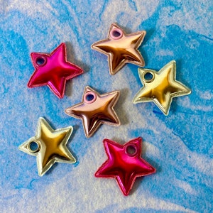 May include: Seven star-shaped charms in shades of pink, gold, and silver, with a metallic sheen. Each charm has a small hole at the top for stringing. The charms are arranged on a blue and white textured surface.