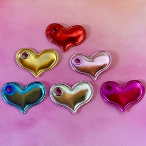 May include: A collection of six heart-shaped charms in various metallic colors: red, gold, silver, pink, blue, and magenta. Each charm has a small hole at the top. The charms are arranged on a pink surface, creating a vibrant and eye-catching display.