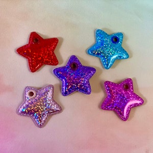 May include: Five glittery star-shaped charms in red, blue, purple, and iridescent silver. Each star has a small hole at the top. The charms are on a gradient background of pink, yellow, and white.