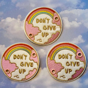 May include: Three embroidered patches with a rainbow, clouds, and the text "DON'T GIVE UP." The patches are circular with a cream-colored background, a gold border, and pink grommets. The rainbow features red, orange, yellow, and blue colors.