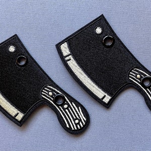 May include: Two black embroidered patches in the shape of a cleaver. Each patch features a white embroidered blade and handle details. The handle has vertical white stripes and a black ring. The patches are on a gray background.