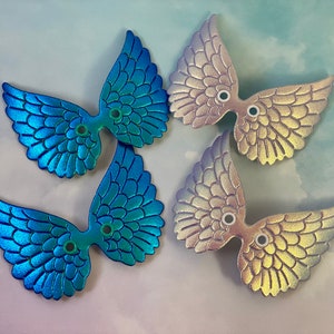 May include: Four iridescent wing-shaped embellishments. Two are blue-green, and two are a mix of pink and gold. Each wing has a detailed feather design and two small holes for attachment. The wings are arranged on a light blue background.