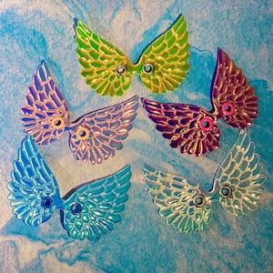 May include: A collection of iridescent, colorful wing-shaped embellishments. The wings are in shades of green, purple, blue, and clear, with a metallic sheen. Each wing has a small, circular hole in the center for attachment.