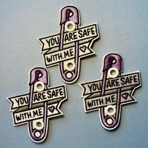 May include: Three embroidered patches in the shape of a safety pin. The patches are purple and white with the text "YOU ARE SAFE WITH ME" in black. Each patch has a small heart symbol.