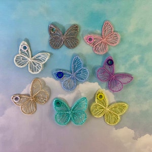 May include: A collection of eight decorative butterfly embellishments in various colors. The butterflies are made of a delicate, openwork material, with a small eyelet in the center of each wing. Colors include white, gray, pink, blue, purple, gold, teal, and yellow.