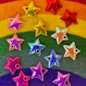 May include: A collection of iridescent star-shaped embellishments in various colors, including yellow, red, pink, purple, and blue. Each star has a small hole for attaching to a project. The stars are arranged on a rainbow-colored background.