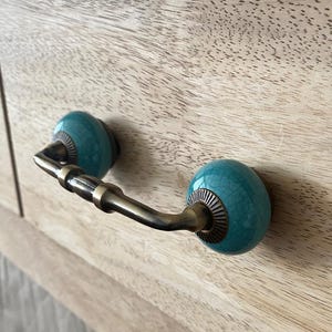 May include: Close-up of a wooden cabinet with a drawer pull. The drawer pull has a brass handle and turquoise ceramic knobs with a crackle glaze. The cabinet is made of light-coloured wood with visible grain.
