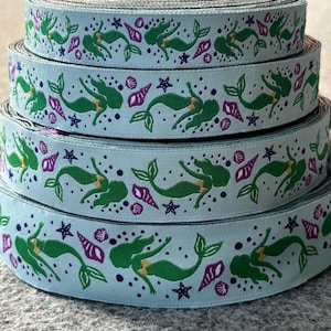 May include: Spools of light blue ribbon with a repeating pattern of green mermaids, purple seashells, and blue starfish. The ribbon is stacked, showing the detailed design. This ribbon is suitable for crafting and embellishments.