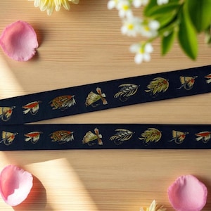 May include: Two strips of dark navy blue fabric ribbon featuring a repeating embroidered pattern of various colorful fishing flies in shades of brown, yellow, green, and orange, arranged in a horizontal line across the length of the material.