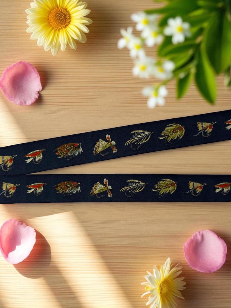 May include: Two strips of dark navy blue fabric ribbon featuring a repeating embroidered pattern of various colorful fishing flies in shades of brown, yellow, green, and orange, arranged in a horizontal line across the length of the material.