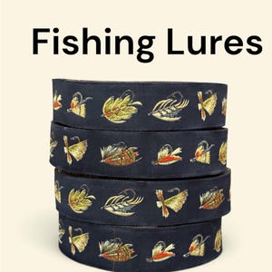 Può includere: Una pila di quattro nastri in tessuto blu navy scuro con motivi ricamati ripetitivi di esche da pesca in tonalità giallo, arancione, verde e marrone. Il testo Fishing Lures è stampato in nero sopra i nastri impilati.