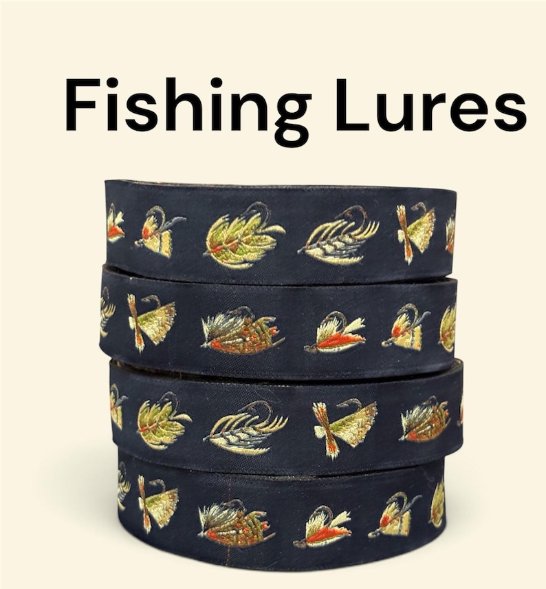 May include: A stack of four dark navy blue fabric ribbons featuring repeating embroidered fishing lure designs in shades of yellow, orange, green, and brown. The text Fishing Lures is printed in black above the stacked ribbons.