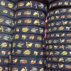May include: A stack of dark navy blue fabric ribbon rolls featuring a repeating pattern of colorful fishing flies in shades of yellow, orange, brown, and white. The ribbon has a smooth texture and is wound into tight, cylindrical spools.