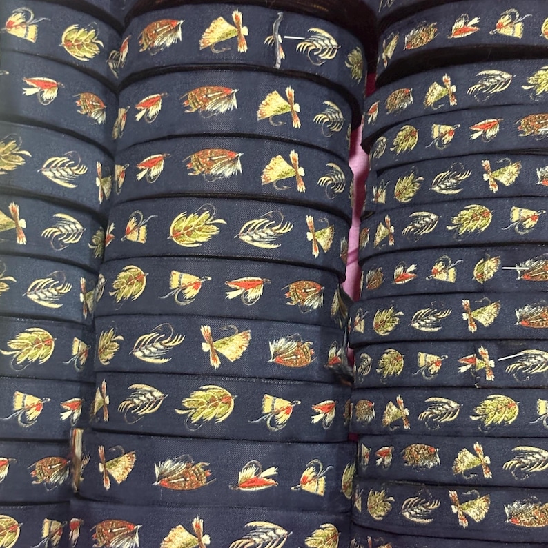 May include: A stack of dark navy blue fabric ribbon rolls featuring a repeating pattern of colorful fishing flies in shades of yellow, orange, brown, and white. The ribbon has a smooth texture and is wound into tight, cylindrical spools.