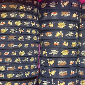 May include: A collection of dark navy blue ribbon spools stacked in columns. Each ribbon features a repeating pattern of colorful fishing flies in shades of yellow, orange, brown, and white, depicting various feathered lure designs.