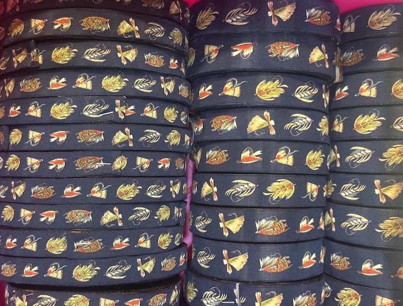 May include: A collection of dark navy blue ribbon spools stacked in columns. Each ribbon features a repeating pattern of colorful fishing flies in shades of yellow, orange, brown, and white, depicting various feathered lure designs.