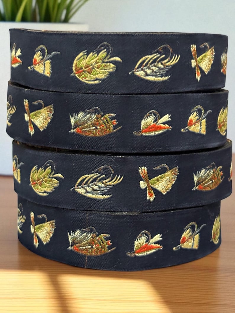 May include: A stack of dark navy blue ribbon featuring an embroidered pattern of various colorful fishing flies. The embroidery includes shades of green, yellow, orange, and white, depicting detailed hooks and feathered lures along the fabric.