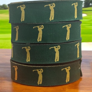 Könnte beinhalten: Gestapelte Rollen dunkelgrünes Band mit einem sich wiederholenden goldenen Golfspieler-Design. Das Band liegt auf einer Holzoberfläche, im Hintergrund ist ein Golfplatz zu sehen.