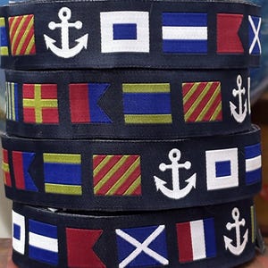 May include: Dark blue nautical ribbon with embroidered signal flags and white anchor designs. The ribbon showcases a repeating pattern of coloured flags, including red, white, blue, and yellow. Ideal for maritime-themed crafts and projects.