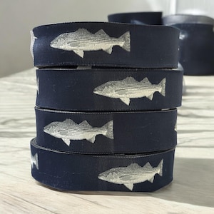 以下が含まれることがあります： 刺繍された銀色の魚が付いた、濃いネイビーブルーのリボンのスプールのスタック。リボンは積み重ねられています。魚のデザインはリボン全体に繰り返されています。背景は明るい色です。