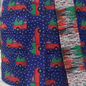 May include: A stack of blue ribbon with a repeating pattern of red trucks carrying green Christmas trees and white polka dots. A separate ribbon with red, silver, and green fringe is visible.