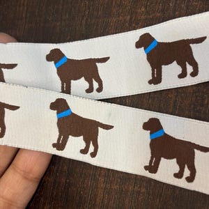 Può includere: Nastro color crema con un motivo ripetuto di Labrador Retriever marroni con collari blu. Il nastro è esposto su uno sfondo di legno scuro, mostrando il design del cane.