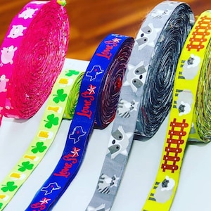 May include: Five different rolls of ribbon with various patterns and colours. The ribbons include a pink ribbon with pigs, a green ribbon with shamrocks, a blue ribbon with the text "Lone Star" and a star, a grey ribbon with raccoons, and a yellow ribbon with sheep and a brown fence.