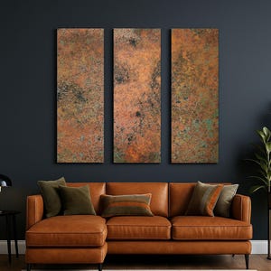 May include: A living room scene featuring a brown leather sectional sofa with several throw pillows. Above the sofa are three vertical rectangular wall art pieces with a textured, copper-colored finish. A black side table with a lamp and a tall plant in a gold pot complete the decor.