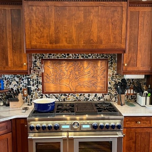 May include: A stainless steel double oven range with a blue pot on the top burner. The backsplash is made of small, round, multi-colored stones with a copper panel featuring a tree design.