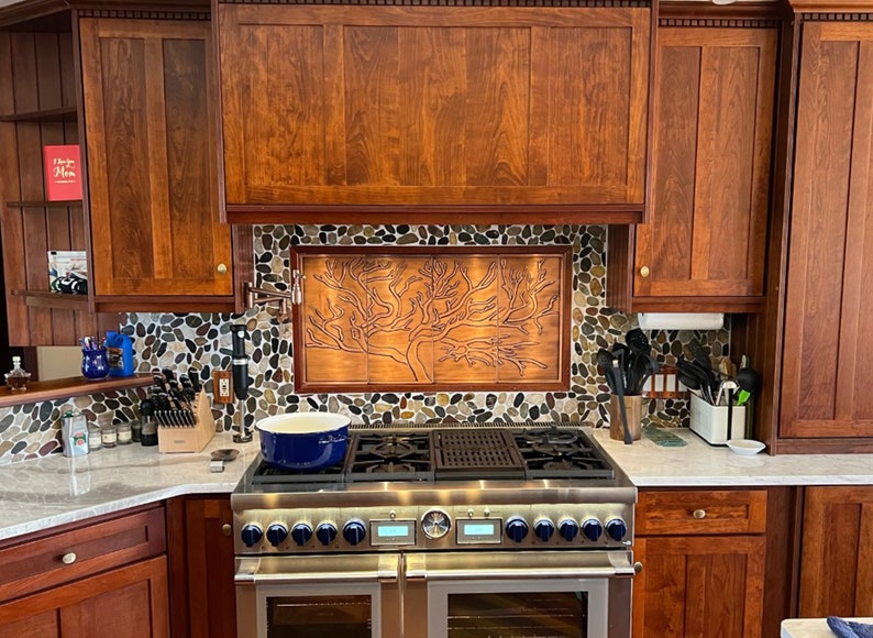 May include: A stainless steel double oven range with a blue pot on the top burner. The backsplash is made of small, round, multi-colored stones with a copper panel featuring a tree design.