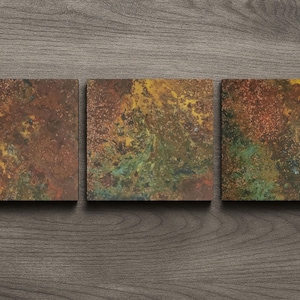 May include: Three square coasters with a textured, abstract design. The coasters feature a blend of colours, including brown, gold, green, and red, creating a rustic appearance. They are arranged on a dark wood surface.