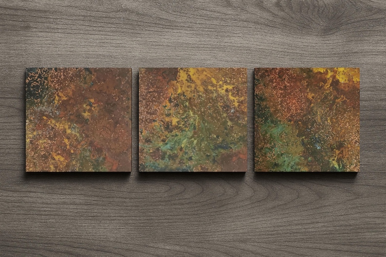May include: Three square coasters with a textured, abstract design. The coasters feature a blend of colours, including brown, gold, green, and red, creating a rustic appearance. They are arranged on a dark wood surface.