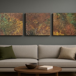 May include: Three square abstract artworks with a textured, mottled appearance in shades of brown, green, and yellow. The art hangs above a light-coloured sofa with olive and tan cushions.