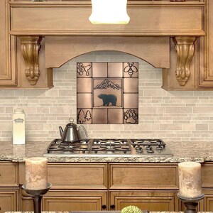 Puede incluir: Un protector de salpicaduras de azulejos de cobre con un dise&ntilde;o de oso, monta&ntilde;a, &aacute;rboles, abeja y huella de pata. El protector de salpicaduras est&aacute; instalado sobre una estufa de gas en una cocina con gabinetes de madera.