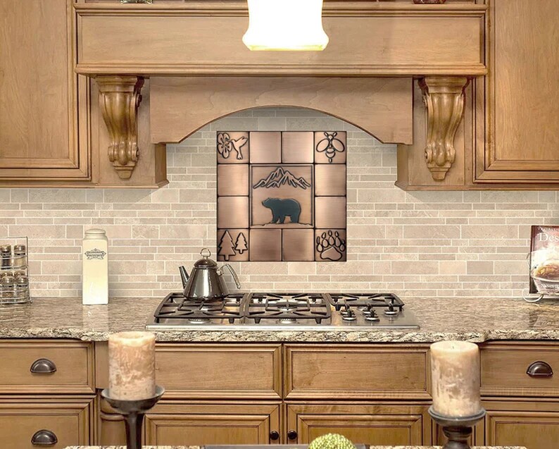 Puede incluir: Un protector de salpicaduras de azulejos de cobre con un dise&ntilde;o de oso, monta&ntilde;a, &aacute;rboles, abeja y huella de pata. El protector de salpicaduras est&aacute; instalado sobre una estufa de gas en una cocina con gabinetes de madera.