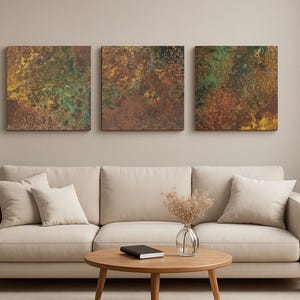 May include: Three square abstract art pieces with textured surfaces in shades of brown, green, and yellow hang above a beige sofa. The room features a round wooden coffee table and neutral-toned decor.