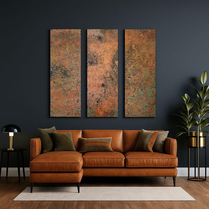 May include: A living room scene featuring a brown leather sectional sofa with several throw pillows. Above the sofa are three vertical rectangular wall art pieces with a textured, copper-colored finish. A black side table with a lamp and a tall plant in a gold pot complete the decor.