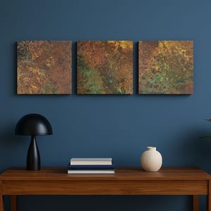 May include: Three square abstract art pieces with a textured, mottled appearance in shades of brown, green, and yellow. The artwork is mounted on a dark blue wall above a wooden table. A black lamp, books, and a white vase are on the table.
