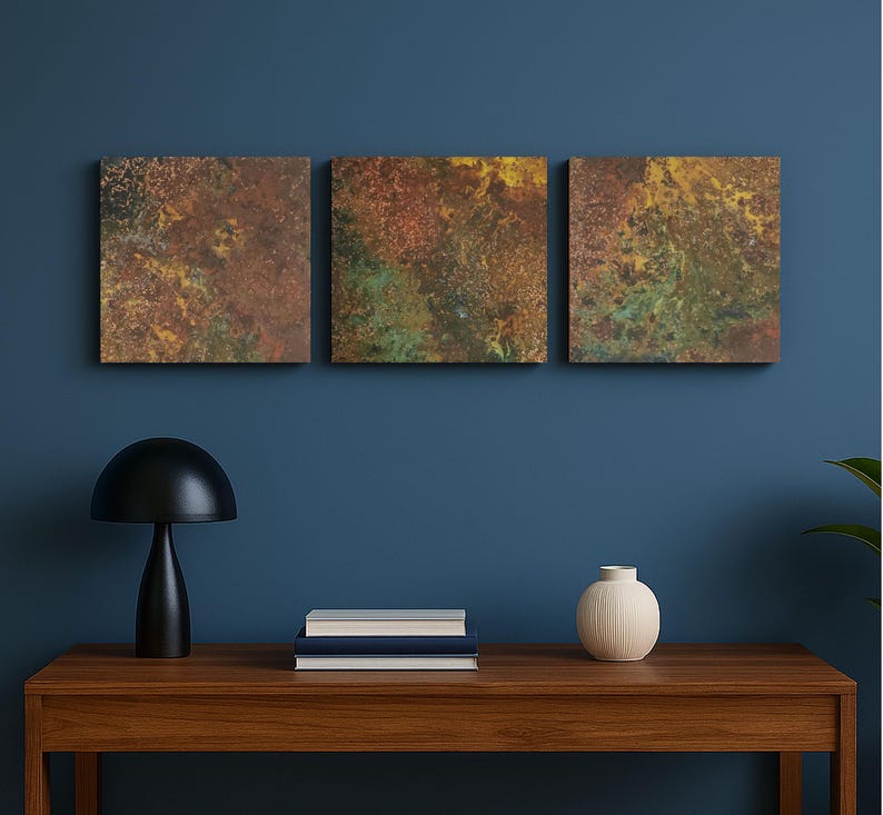 May include: Three square abstract art pieces with a textured, mottled appearance in shades of brown, green, and yellow. The artwork is mounted on a dark blue wall above a wooden table. A black lamp, books, and a white vase are on the table.
