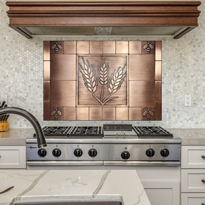 Pode incluir: Uma placa de metal com tom de cobre com um design de espiga de trigo acima de um fogão a gás de aço inoxidável. A placa é emoldurada com detalhes decorativos de abelhas. A cozinha apresenta armários brancos e uma bancada de mármore.