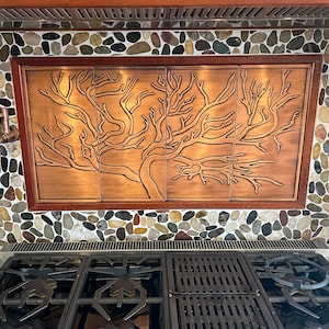 May include: A copper kitchen backsplash with a tree design. The backsplash is installed above a stainless steel gas stove with black grates.