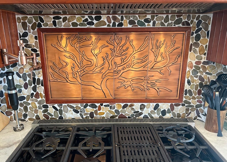 May include: A copper kitchen backsplash with a tree design. The backsplash is installed above a stainless steel gas stove with black grates.