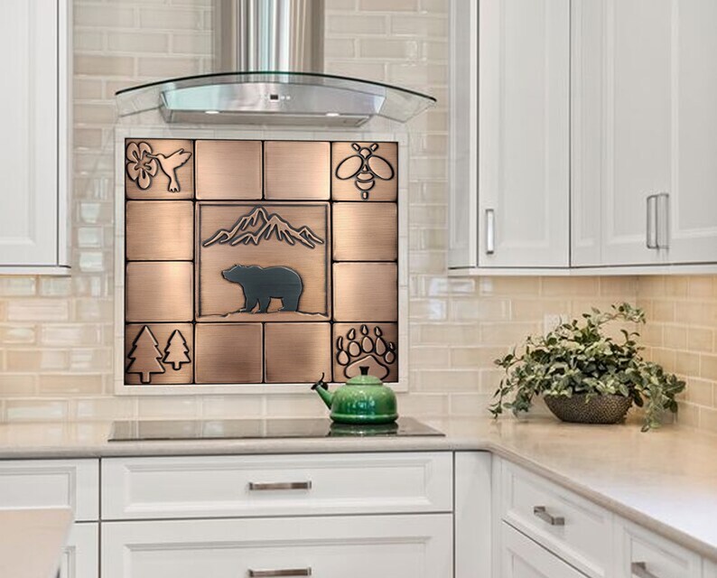 Puede incluir: Un salpicadero de azulejos de color cobre con un oso, monta&ntilde;as, &aacute;rboles, una abeja, una huella de pata y un colibr&iacute;. El salpicadero est&aacute; instalado sobre una encimera de cocina blanca.