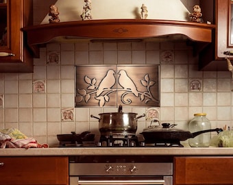 May include: A kitchen backsplash featuring two rectangular metal tiles with a bronze finish, depicting two birds perched on a branch. The tiles are centered above a stovetop with a stainless steel pot and a black skillet.