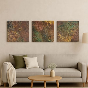 May include: Three square abstract artworks with a textured, mottled appearance. The colour palette includes shades of brown, green, and yellow. The artworks are displayed above a light grey sofa with decorative cushions and a wooden coffee table.