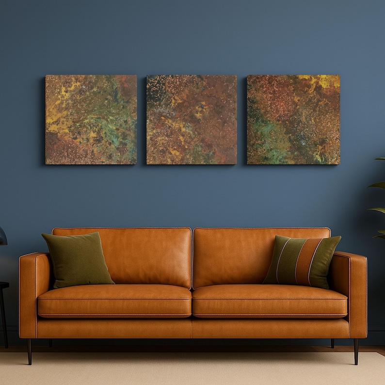 May include: Three square abstract art pieces with a textured, mottled appearance in shades of brown, green, and yellow are mounted on a dark blue wall. Below, a brown leather sofa with two green cushions.